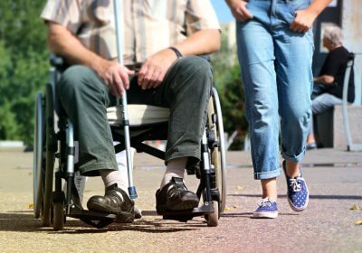 Muška osoba u invalidskim kolicima sa štapom i druga ženska osoba iza nje