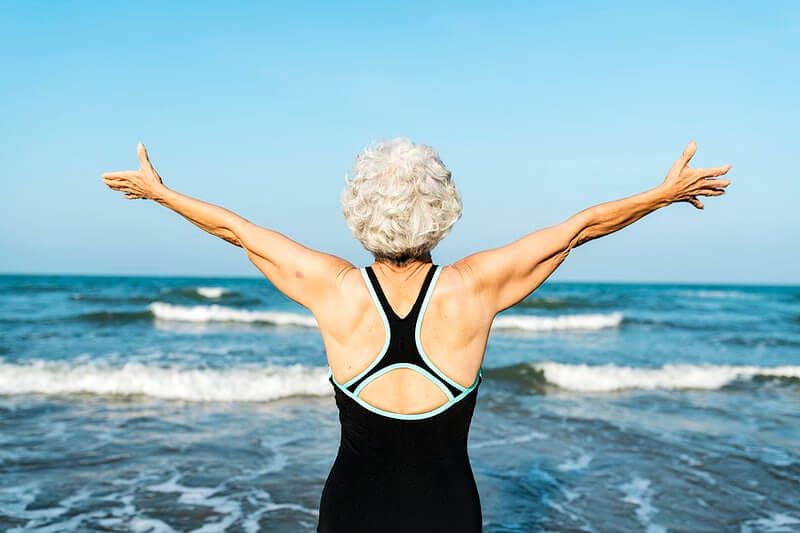 Starija ženska osoba u kupaćem kostimu raširenih ruku stoji ispred mora