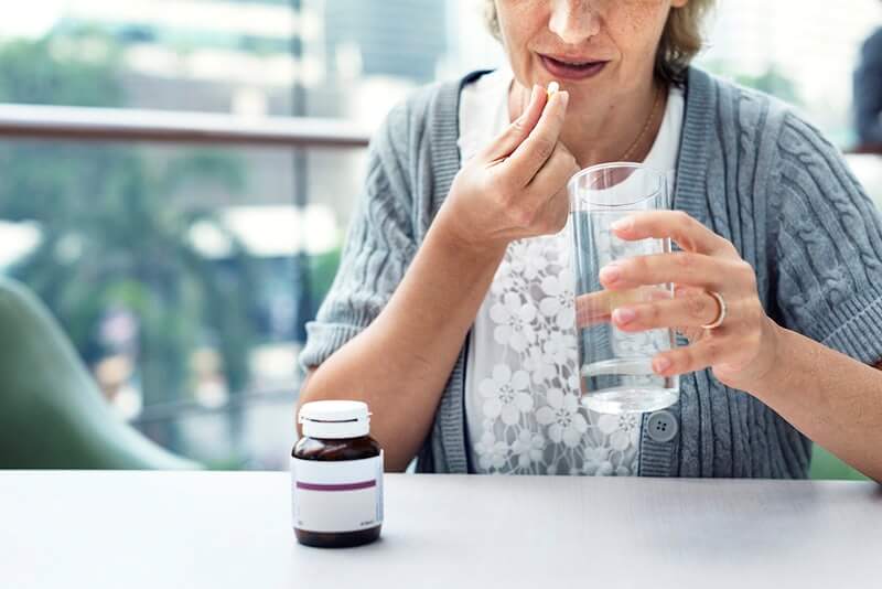 Starija gospođa uzima lijek s jednom rukom, a u drugoj drži čašu vode