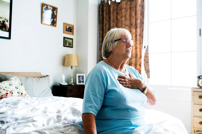 Starija ženska osoba se drži za srce dok sjedi na krevetu