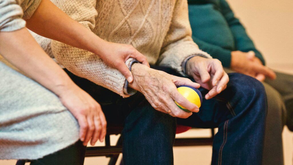 osoba u domu za starije drži ruku umirovljenika koji u ruci ima lopticu i oboje sjede