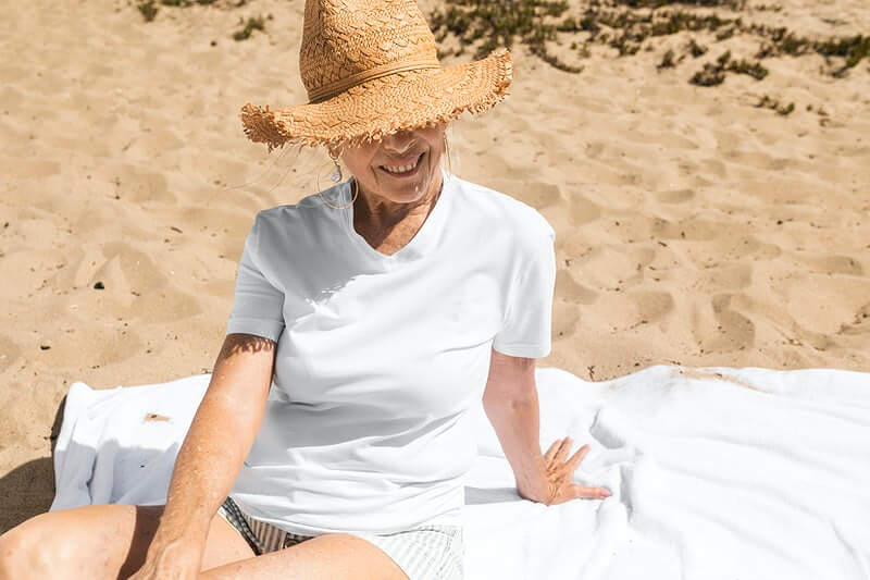 Starija žena sjedi na plaži u bijeloj majici i šeširom na glavi