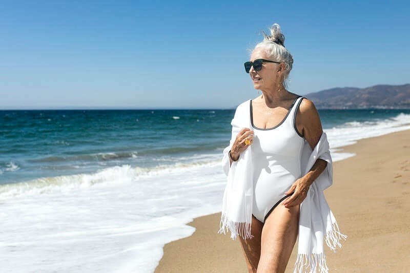 Starija gospođa u bijelom kupaćem kostimu i bijeloj košulji na plaži