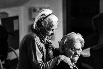 Crno-bijela fotografija dvije umirovljenice, pri čemu jedna stoji i drži ruku na ramenu umirovljenice koja sjedi