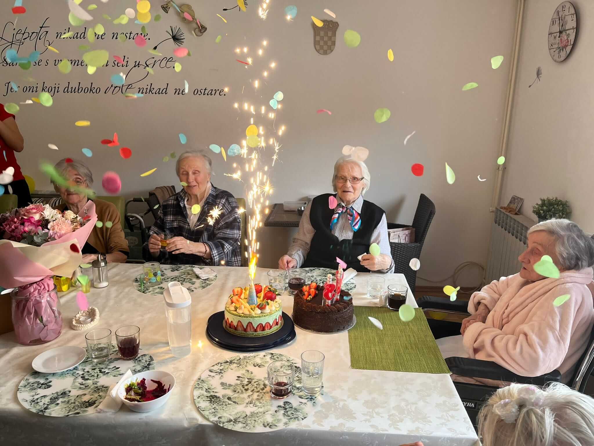 proslava rođendana četiri umirovljenice u domu za starije osobe s dvije torte na stolu, svjećicama koje gore i konfetima u zraku