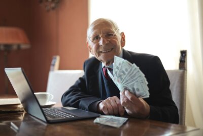 Umirovljenik u odijelu sjedi s novcima u rukama, a ispred njega je na stolu laptop