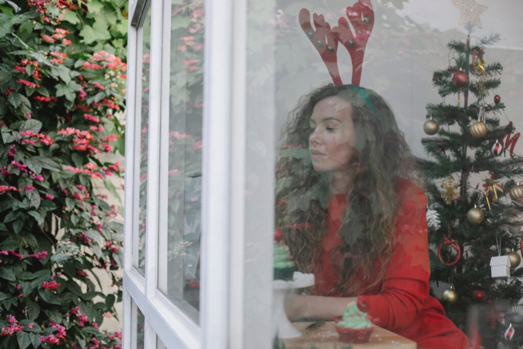 Zamišljena žena sjedi u sobi s božićnim drvcem i ukrasima dok gleda kroz prozor