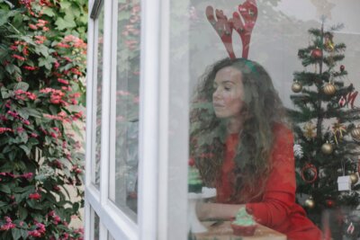 Zamišljena žena sjedi u sobi s božićnim drvcem i ukrasima dok gleda kroz prozor
