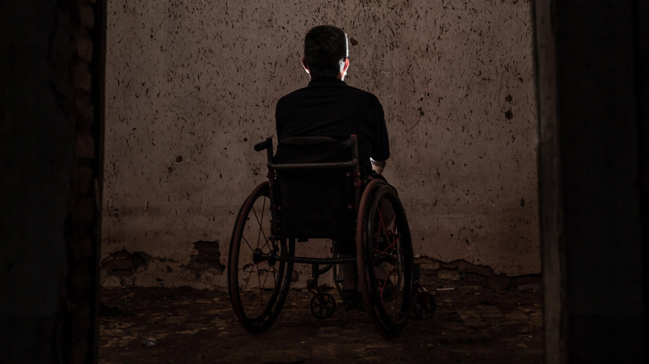 Osoba s invaliditetom u invalidskim kolicima sjedi u tami okrenuta prema zidu
