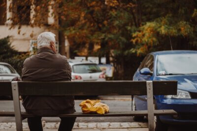 Sijedi umirovljenik u smeđoj jakni sjedi na klupi.