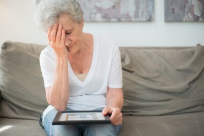 Umirovljenca u bijeloj kratkoj majici sjedi na kauču s tabletom u krilu i drži se za glavu