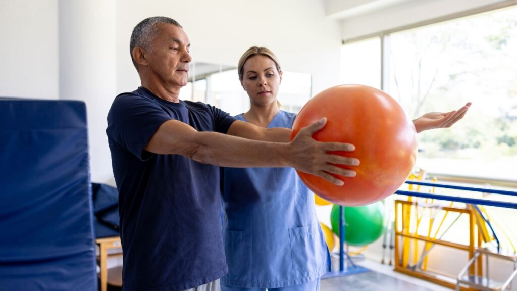 Stariji muškarac na fizioterapiji drži narančastu loptu u ruci