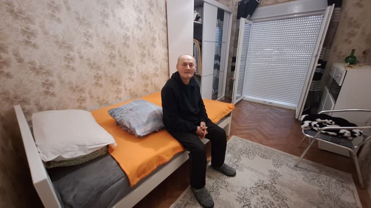 Nisvet sjedi na krevetu u sobi s tepihom i ormarom