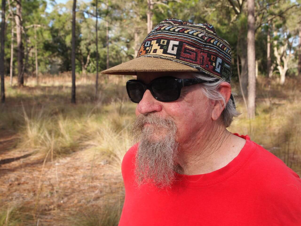 Umirovljenik u crvenoj majici sa sunačnim naočalama i šarenom šiltericom vani u prirodi